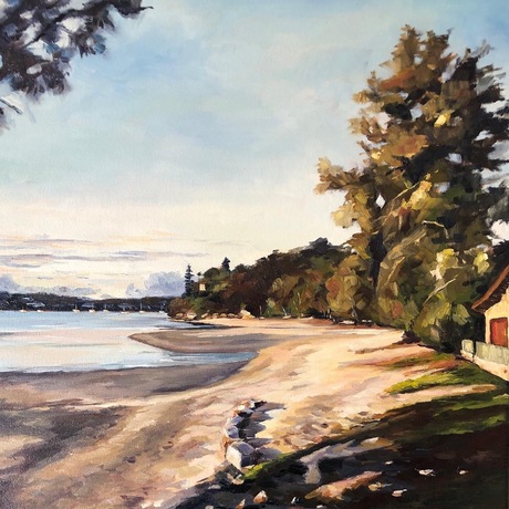 A beach scene of a bay at sunset as last light results in  golden light and long shadows. There are trees along side the sand with sunlight hitting the trees. Headland and boats in the the distance.  