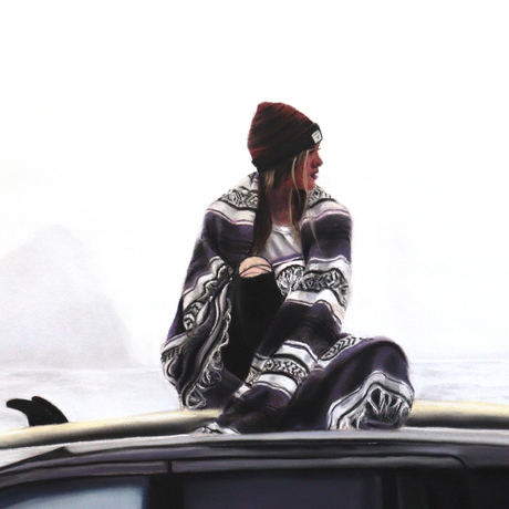 girl wrapped in blanket sitting on surfboard on top of a car in front of ocean on a winter day