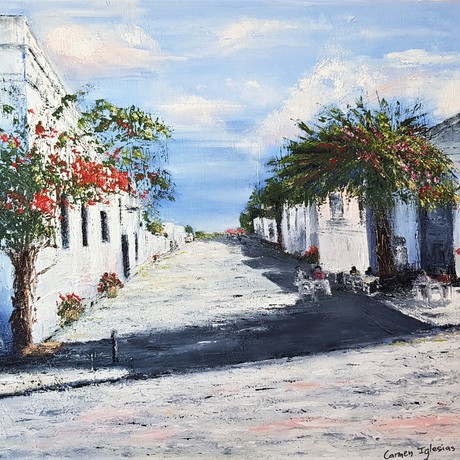 View of one of the many little streets in Colonia del Sacramento, Uruguay. Historic white washed houses border the narrow street which ends on the River Plate in the background.  A small cafe on one of the corners with tables and chairs on the footpath and people sitting, enjoying the sun. Trees covered in red and white flowers on both sides of the streets. Also, pots with red flowers on the sides of the street. 