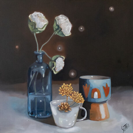 A blue glass bottle on the left with three poppy bud stems, a white teacup with a yellow succulent and a blue hand made ceramic votive. Five fireflies or orbs sparkling in the still life scene