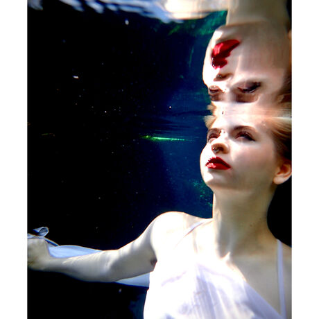 A surreal underwater scene where a girl in a white dress, who has light skin and blonde hair, is partially submerged in water and looks upon her reflection.