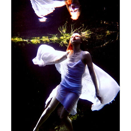 An ethereal underwater scene where a girl in a white dress, swims through the water and looks upon her reflection.