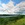 Impressionist rendering of bright clouds over a lake and mountains on a summer day, with green hills and a grassy foreground.