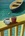 Two mix matched coffee cups rest on a yellow hand rail over looking turquoise water and a small row boat resting on a mooring.