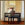 A young boy about the age of 7 sits at a piano and practices with his back turned to us. He is framed on both sides by drapes illuminated with outside daylight.