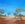 The image shows a dry colourful landscape with some trees. Father Emu takes his children to a small waterhole. 