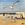 A seagull flies in the foreground of a nostalgic beach scene with ice cream van, paper boy and beachgoer in a bikini.