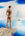 A nude male figure stands high above the ocean on top of a cliff face as he looks out into the distance. Above, the sky is a deep blue, mostly filled with white clouds. The ocean has patches of light and deep blues. Beyond is a single island of rock.