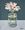 A small glass vase with white daisies on a background split horizontally in two eith light blue on the bottom third and dark blue on the top 2 thirds.  There are light reflections on the glass vase.