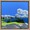 Rocky foreground before bright green grass, trees and ocean  with distant buildings and trees under a cloudy sky.