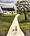 Countryside scene with a white cottage with a black slate roof set up high at the end of a long white road. There is a lady wearing an orange and black striped dress walking her black dog on the white road. The road leads up to the white cottage. On each side of the road there is green grass ranging from light olive green to dark olive green. On the right side there is a large tree. The sky is filled with grey clouds. The bottom foreground has wisps of lilac purple wildflowers. 