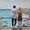 A young couple looking out to sea. He with dark hair, wearing  a white linen shirt. She with blonde hair and a warm brown dress.