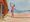 artwork showing a man on the beach dressed in swim shorts walking the tight rope tied to the wooden pier 
