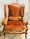 A velvet, rust coloured french provincial chair in the sunlight on a parquetry style floor, with a Provencal blue french door behind