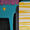 A playful modern composition featuring a turquoise blue background with pink scalloped edges along the top. A vibrant yellow circle, suggesting a sun, floats in the turquoise space. The right side of the canvas is divided by bold yellow and white horizontal stripes, creating a rhythmic pattern. Dark geometric shapes in black and grey add depth and structure to the left side. The overall effect is cheerful and contemporary, with the bright color palette living up to its title 'Sunny colour mood'. 