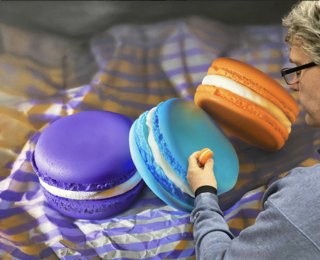 painting of three beautifully made macrons on a purple  and brown striped paper sheet. The sheet is crumpled so it adds texture and shadow. The three macrons are resting on each other, a purple , a bright peacock blue and an bright orange one all vying for attention of the viewer.  the background is dark and in shadow to create a dramatic backdrop for the bright macrons.  the painting looks realistic but of course it is a thousand times bigger than real size!  the painting looks like a photo but if you look closely, you can see the almost abstract brushstrokes that create the detail!