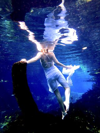 Young woman standing on a log underwater in white dress whilst reaching her foot back en pointe.