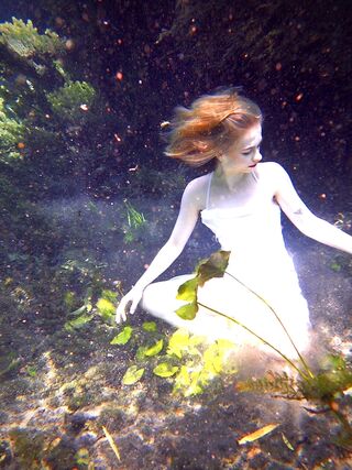 Young woman underwater in white dress sitting amongst a sea garden.