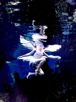 Young woman sitting upon a log underwater in white dress with fairy wings - patiently waiting.