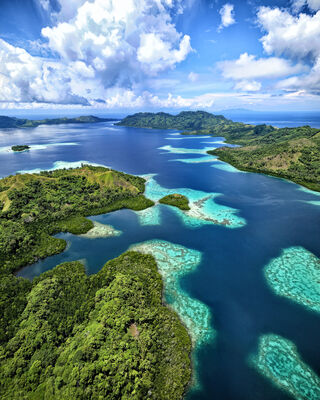 Forested islands surround a deep blue bay with isolated turquoise coloured coral reefs. Billowing monsson clouds fill the sky.