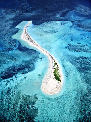 A large swirling shaped sand bar in turquoise blue seawater with emerald green patches of vegetation on the white sand, surrounded by dark blue-black seagrass beds.