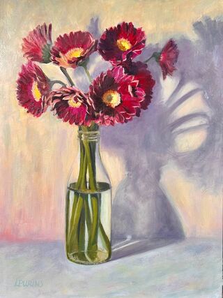 A bunch of deep red burgundy gerberas with yellow to orange circular centres in a glass bottle with water on a pale peach and aqua background. The light is also creating a large shadow behind the flower arrangement which is part of the composition. 