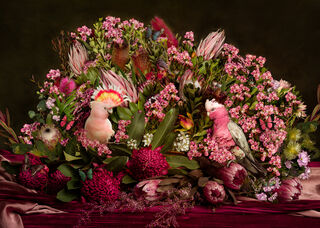 Large bunch of colourful native Australian flowers arranged on a table. The signature colour is pink. Inside the arrangement are a pink cockatoo and galah. Many butterflies and beetles are hidden in the foliage. 