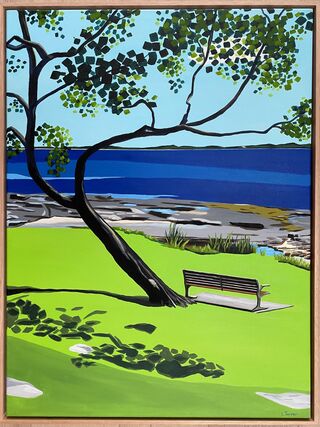 View of an empty bench under a large tree with rocks and ocean in the background. The sky is bright blue and there are dark shadows on the grass in the foreground from the tree.