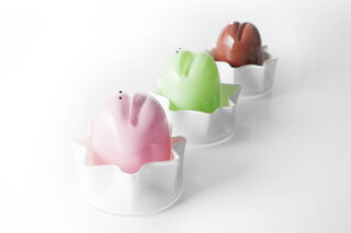 Three glass sculptures resembling frog cakes. Pink, green and brown rounded tubes with mouths and eyes sitting inside a frilly white bowl/vessel resembling the patty pans of a cupcake. 