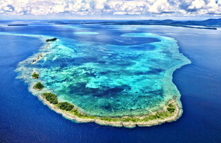 A narrow band of an island surrounds  a turquoise coloured lagoon filled with complex coral reefs, surrounded in turn by  the deep blue sea. Larger islands lie in the distance, stretching to a horizon edged with monsoon clouds.