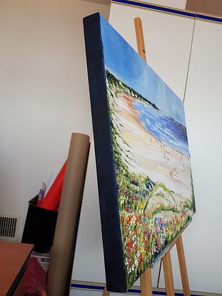 Long and wide beach. People walking on the sand. People sitting under umbrellas. Others sitting on the sand. Greenery and wild flowers on the foreground. Hills in the horizon. 