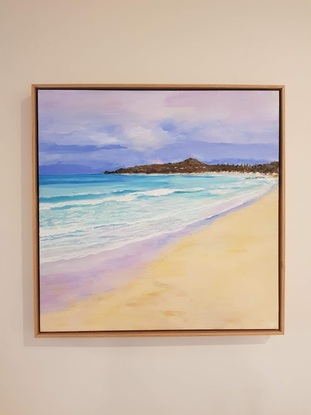 A view across sand with turquoise water and waves. There are small inviting waves and distant deep blue water.  The tiny lighthouse is in the background set against a darkish sky with white clouds and the peninsula at dusk.