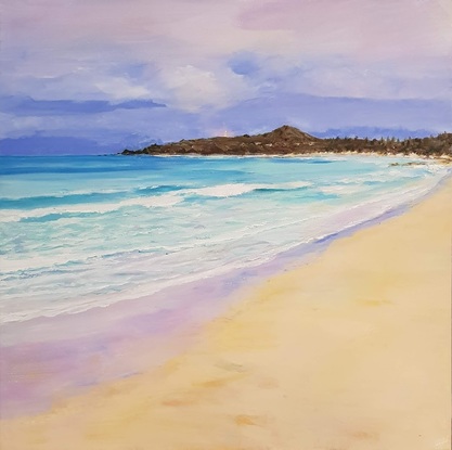 A view across sand with turquoise water and waves. There are small inviting waves and distant deep blue water.  The tiny lighthouse is in the background set against a darkish sky with white clouds and the peninsula at dusk.