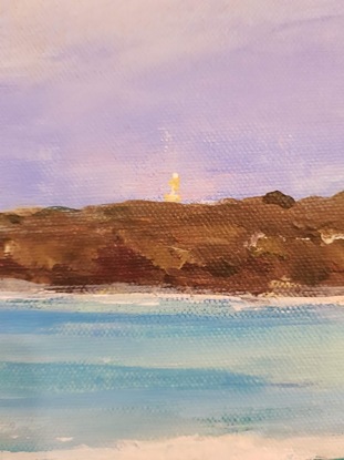 A view across sand with turquoise water and waves. There are small inviting waves and distant deep blue water.  The tiny lighthouse is in the background set against a darkish sky with white clouds and the peninsula at dusk.