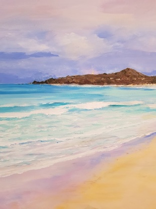 A view across sand with turquoise water and waves. There are small inviting waves and distant deep blue water.  The tiny lighthouse is in the background set against a darkish sky with white clouds and the peninsula at dusk.