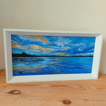 Moody cloudy sunset skies in blues, whites, oranges and yellows, above a calm river reflecting these colours and deeper shades of the same hues. A thin horizon of land is horizontally silhouetted across the centre of the scene.