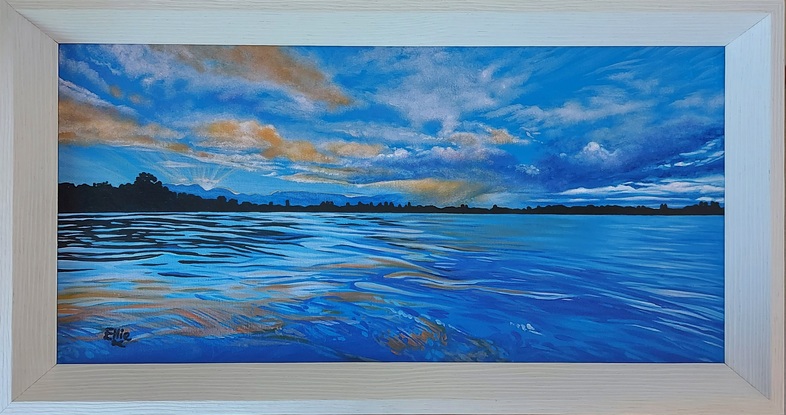 Moody cloudy sunset skies in blues, whites, oranges and yellows, above a calm river reflecting these colours and deeper shades of the same hues. A thin horizon of land is horizontally silhouetted across the centre of the scene.