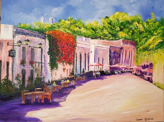 Restaurants and cafes  in heritage buildings with pink and purple colour walls in the Old Town in Colonia del Sacramento, un Uruguay South America.  A sunny day. Tables with red umbrellas on the footpath. People sitting at the tables. A cascade of red flowers covers most of one building. Green trees above and behind the buildings.  Two church towers among the trees above and behind the buildings.  The buildings cast a purple shade on the otherwise sunny road.  