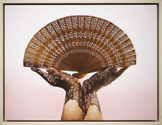 Two hands holding a handheld wooden fan with light streaming through the cut-out patterns of the fan, creating dappled patterns on the two hands. The background is a soft pink.