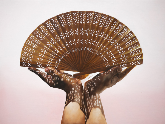 Two hands holding a handheld wooden fan with light streaming through the cut-out patterns of the fan, creating dappled patterns on the two hands. The background is a soft pink.