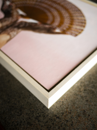 Two hands holding a handheld wooden fan with light streaming through the cut-out patterns of the fan, creating dappled patterns on the two hands. The background is a soft pink.