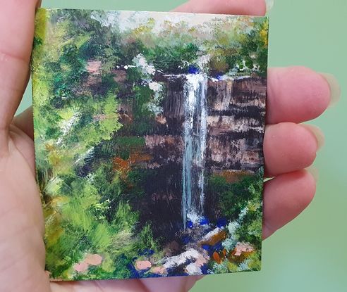 Miniature painting of a small waterfall viewed through a break in green forest; acrylic on stone tile by Victoria Collins.