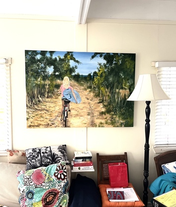 Painting called The Way Home by Banx, of a blond girl on a bicycle carrying a surfboard.