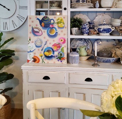 An aerial view of a table set for afternoon tea with cream floral tablecloth blue cups and saucers and a blue and green teapot.  A glass sugarbowl and variety of flowers in vases.