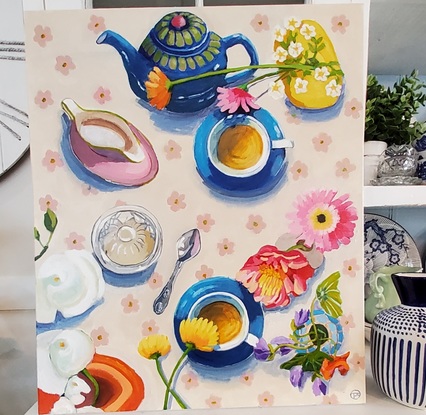 An aerial view of a table set for afternoon tea with cream floral tablecloth blue cups and saucers and a blue and green teapot.  A glass sugarbowl and variety of flowers in vases.