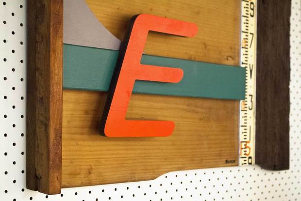 A three dimensional wall sculpture made of reclaimed timber and found objects. The colours in the work are natural wood and red and green. There is a large letter "E" in an orange/red and an aluminium section with a measurement scale on it.