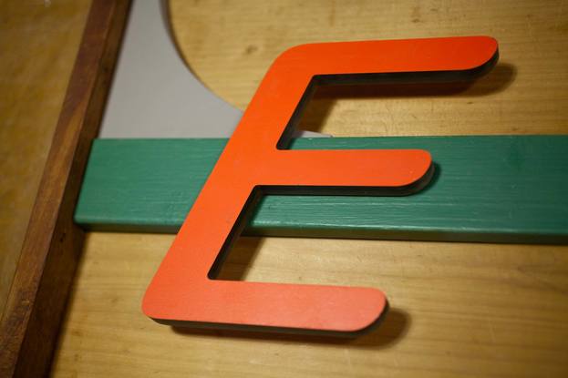 A three dimensional wall sculpture made of reclaimed timber and found objects. The colours in the work are natural wood and red and green. There is a large letter "E" in an orange/red and an aluminium section with a measurement scale on it.