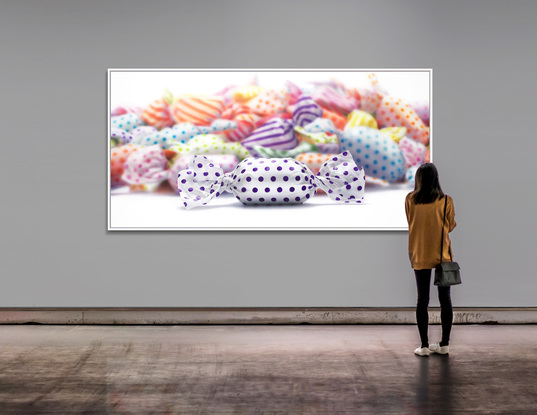 A 2m by 1m painting on canvas of a pile of candy/lolloes in colourful wrappers painted blurred to put attention to one purple polka dotted candy.
The candies are created in hyper realism style -- sharp and in focus. The candies are  a 1,000 times their actual size! .