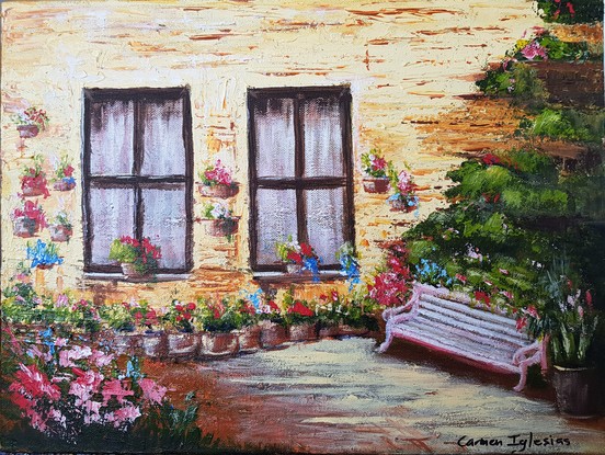 A pinkish/white park bench among colourful flowers. The bench is near an ochre colour wall on which you can see two large windows with white voile curtains and dark brown frames. Several pots with flowers hang from the wall. 