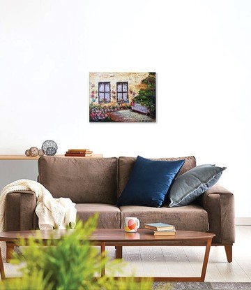 A pinkish/white park bench among colourful flowers. The bench is near an ochre colour wall on which you can see two large windows with white voile curtains and dark brown frames. Several pots with flowers hang from the wall. 